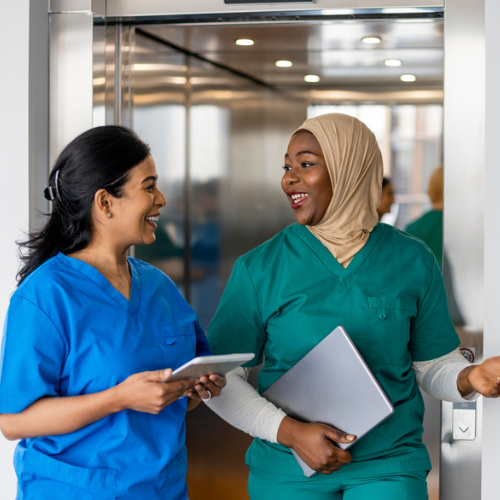 two doctors walking exiting an elevator one with long black hair pulled back with hair clip and one with head wrap both wearing scrubs