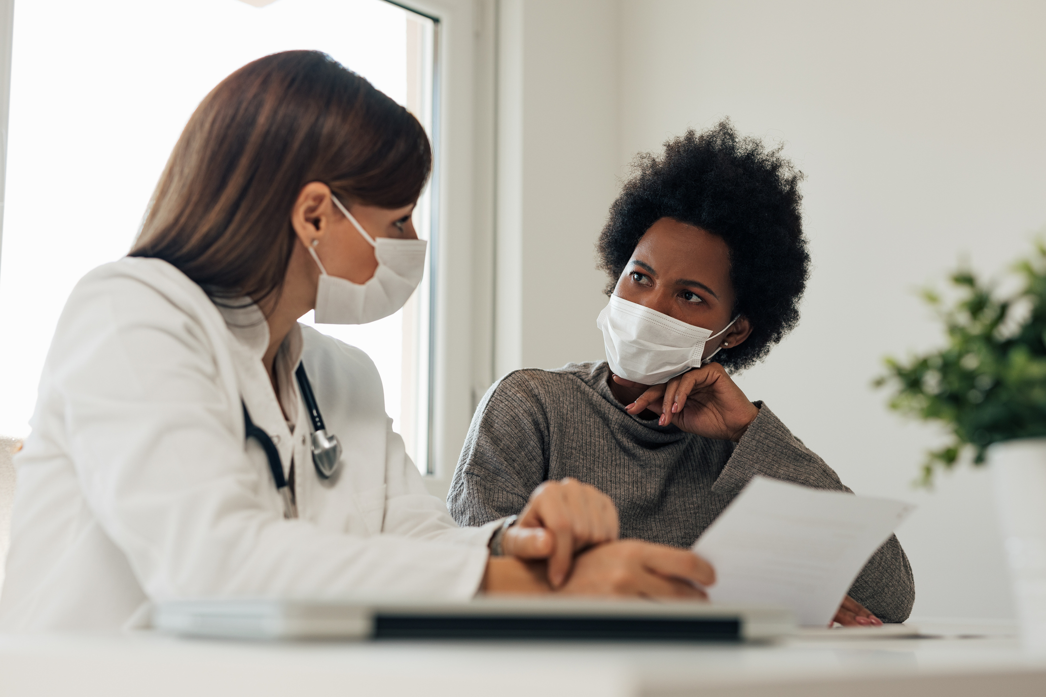 Black person with short afro hair and long sleeve green blouse and mask over mouth talks to doctor with mask