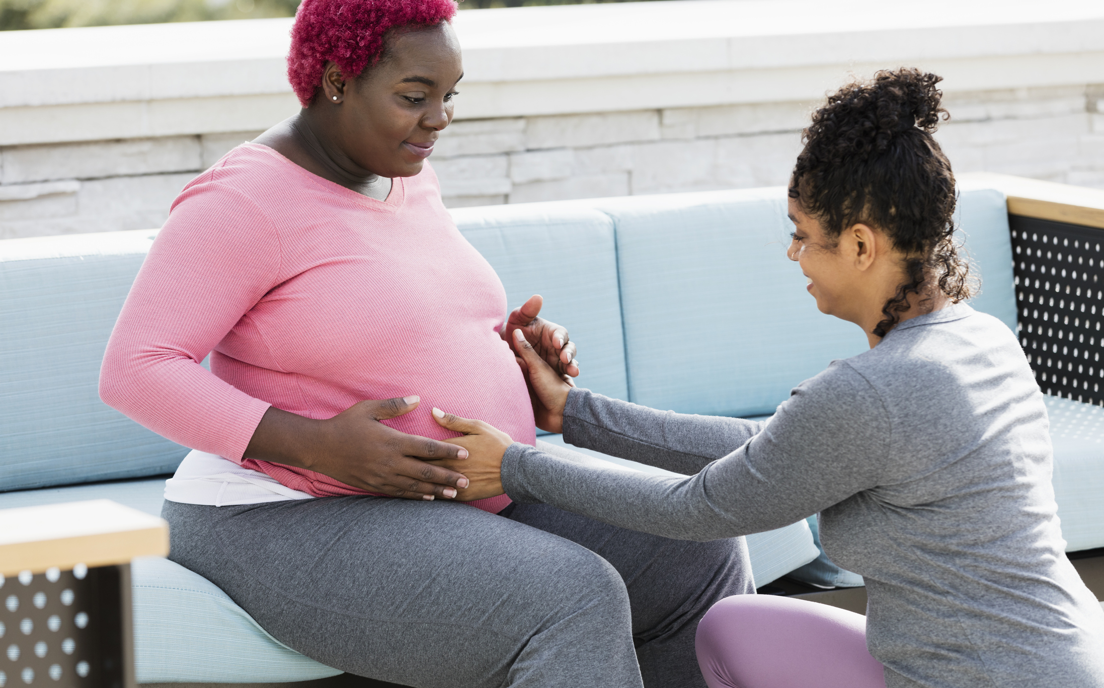 Doula squatting down with long tightly coiled black hair pulled up in a bun, massages belly of Black pregnant person with short hot pink hair sitting on a ball
