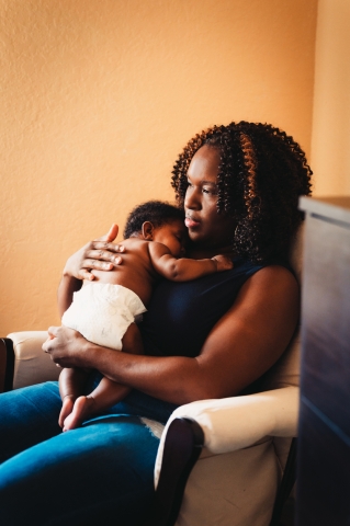 Black mother with curly hair sitting in chair holding baby in diaper resting head on shoulder