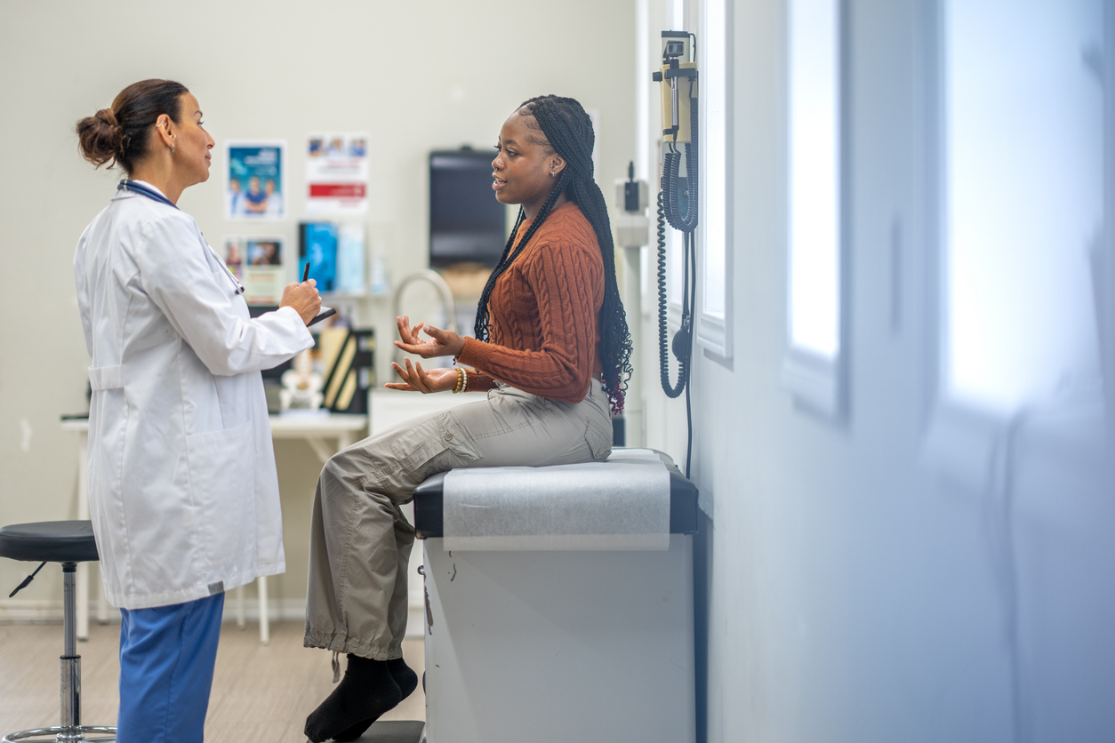 Young teenage Black patient sits on hospital exam chair while talking to female provider