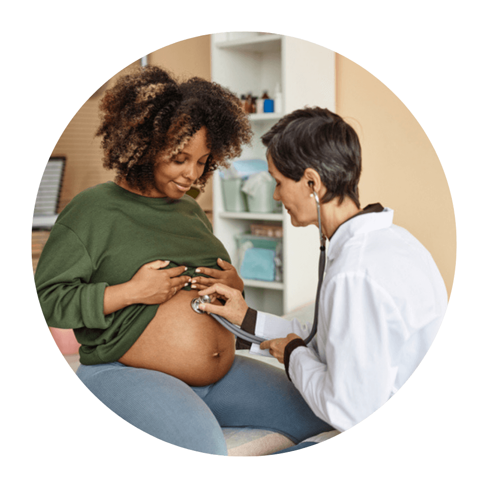 Black pregnant person with curly afro sits on exam table while doctor listens to her belly with her long sleeved green blouse pulled up