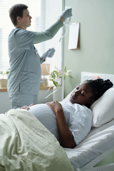 Medical professional attending to Black pregnant woman lying in hospital bed adjusting intravenous drip in medical setting with clipboard on wall and plants on windowsill